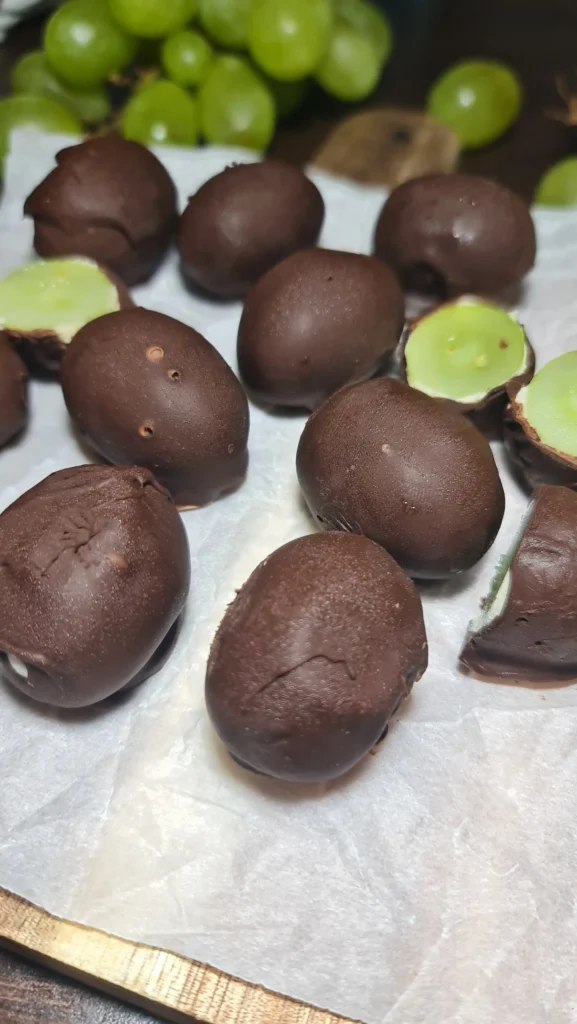 An overhead view of a wooden cutting board with chocolate grape bark bites, surrounded by half a coconut and pineapple slices on a dark wood table.