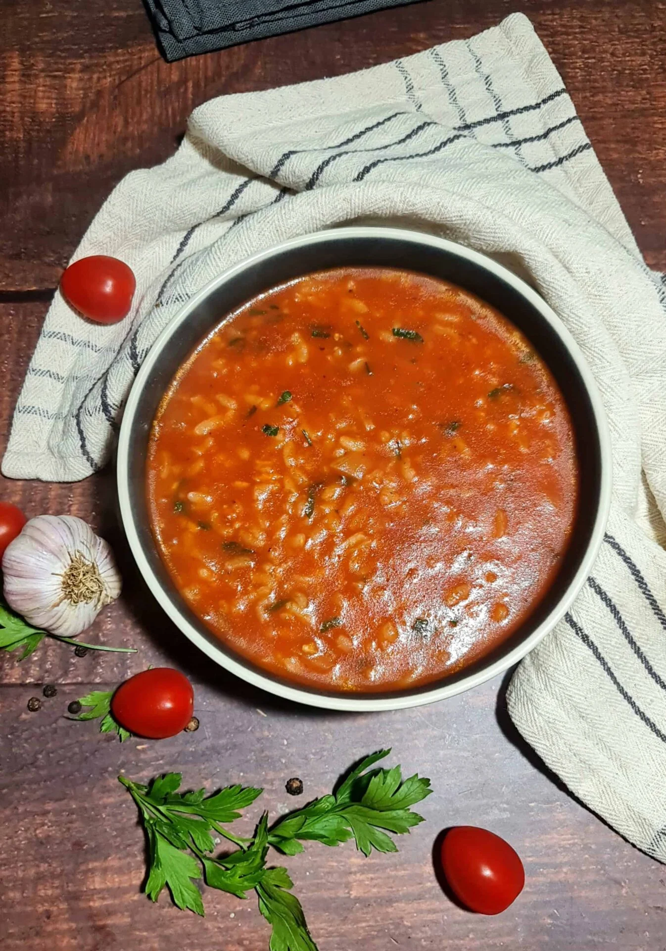 A rustic bowl of thick and hearty tomato rice soup, styled on a dark wood table with a kitchen towel, fresh tomatoes, and garlic.-1