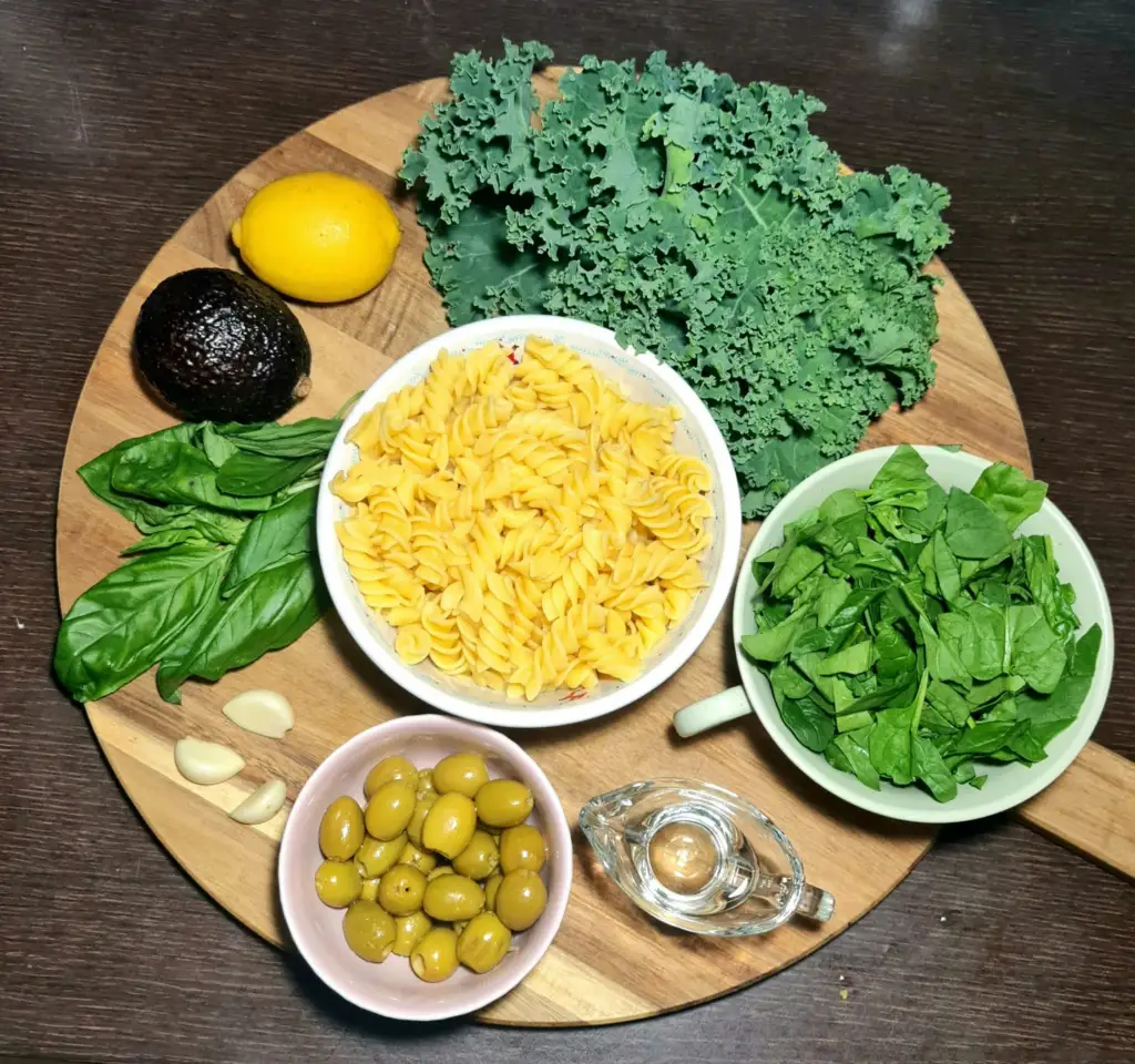 Raw ingredients for making gluten-free avocado green pasta, including raw fusilli, fresh spinach, kale, and lemon on a wooden board.