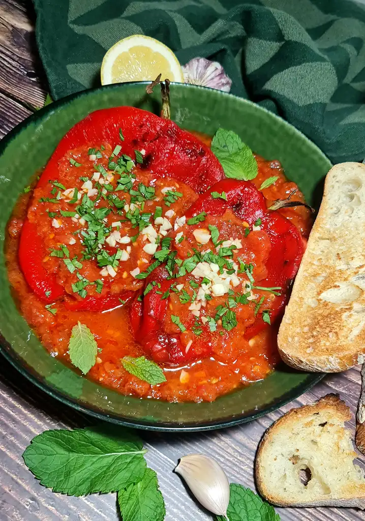 Close-up of roasted red peppers with fresh tomato sauce topped with garlic and parsley, served in a rustic green bowl with crispy bread on the side.