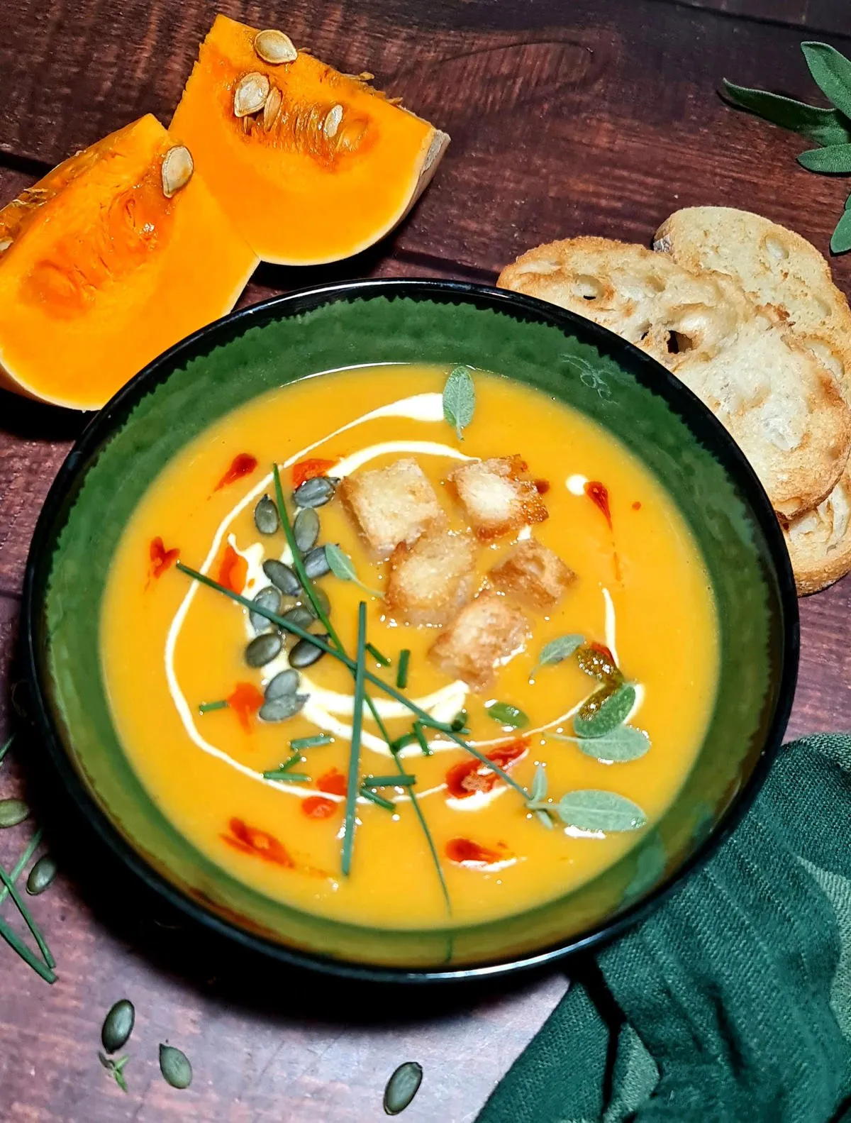 Overhead view of vibrant orange pumpkin soup in dark green ceramic bowl, topped with homemade croutons, roasted pumpkin seeds, fresh herbs, and decorative oil drizzle on dark wood table