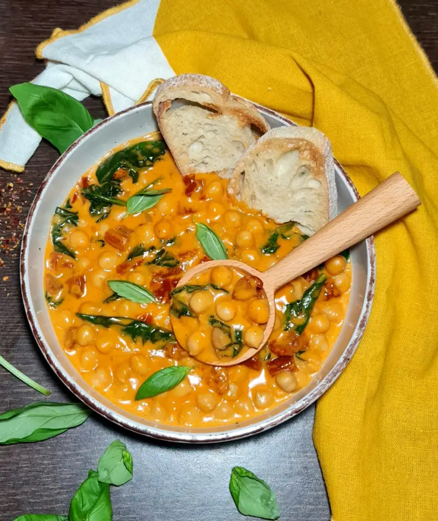 A wooden spoon scooping up a hearty portion of creamy Merry me chickpeas from a garnished bowl with crusty bread.