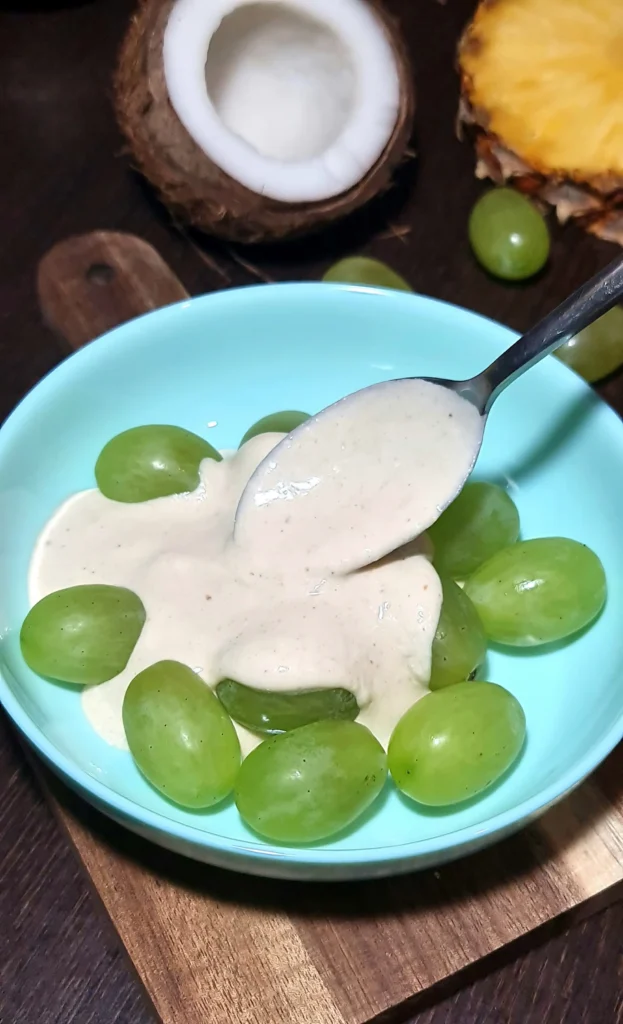 Coating green grapes with a creamy mixture in a light blue bowl for making chocolate grape bark, with coconut in the background.