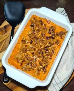 Overhead view of a baked sweet potato casserole in a white rectangular dish, topped with crunchy pecan streusel, next to a black spatula and cinnamon sticks.