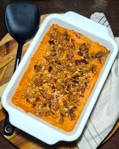 Overhead view of a baked sweet potato casserole in a white rectangular dish, topped with crunchy pecan streusel, next to a black spatula and cinnamon sticks.