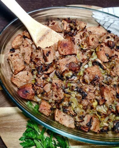 A glass baking dish filled with golden-brown baked vegan stuffing, with a wooden spoon resting inside.