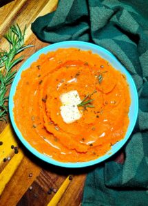A light blue bowl filled with bright orange vegan mashed sweet potatoes, garnished with melting butter, fresh rosemary, and cracked pepper.