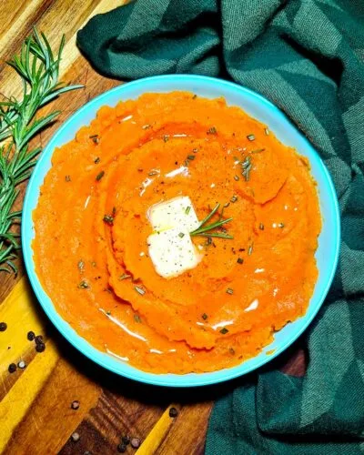 A light blue bowl filled with bright orange vegan mashed sweet potatoes, garnished with melting butter, fresh rosemary, and cracked pepper.