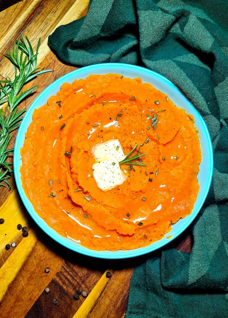 A light blue bowl filled with bright orange vegan mashed sweet potatoes, garnished with melting butter, fresh rosemary, and cracked pepper.