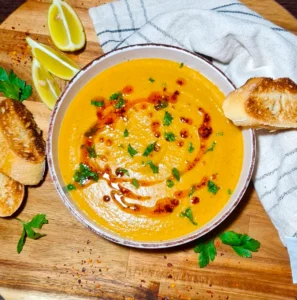 Overhead view of a white bowl filled with golden-yellow Turkish lentil soup, drizzled with red chili oil and chopped parsley, served with toasted bread and lemon wedges on a wooden board.