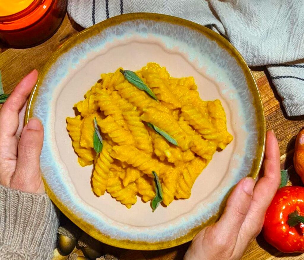 Hands holding a cozy bowl of Creamy Vegan Pumpkin Pasta, garnished with sage, next to a lit candle and decorative pumpkins.