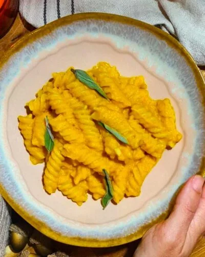 Hands holding a cozy bowl of Creamy Vegan Pumpkin Pasta, garnished with sage, next to a lit candle and decorative pumpkins.
