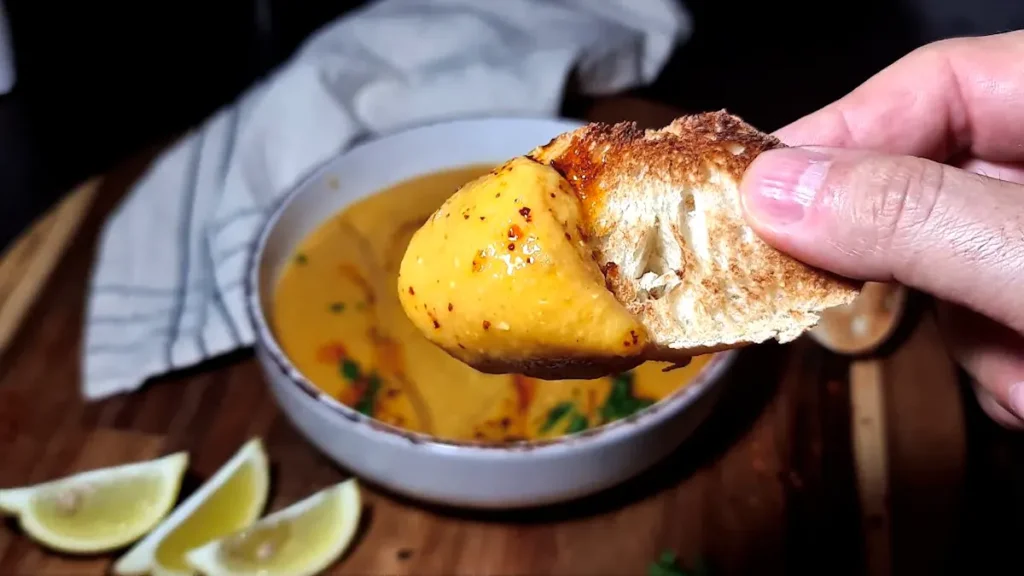 A hand holding a slice of toasted crusty bread dipped into thick, creamy Turkish lentil soup, with the soup bowl and lemon wedges blurred in the background.