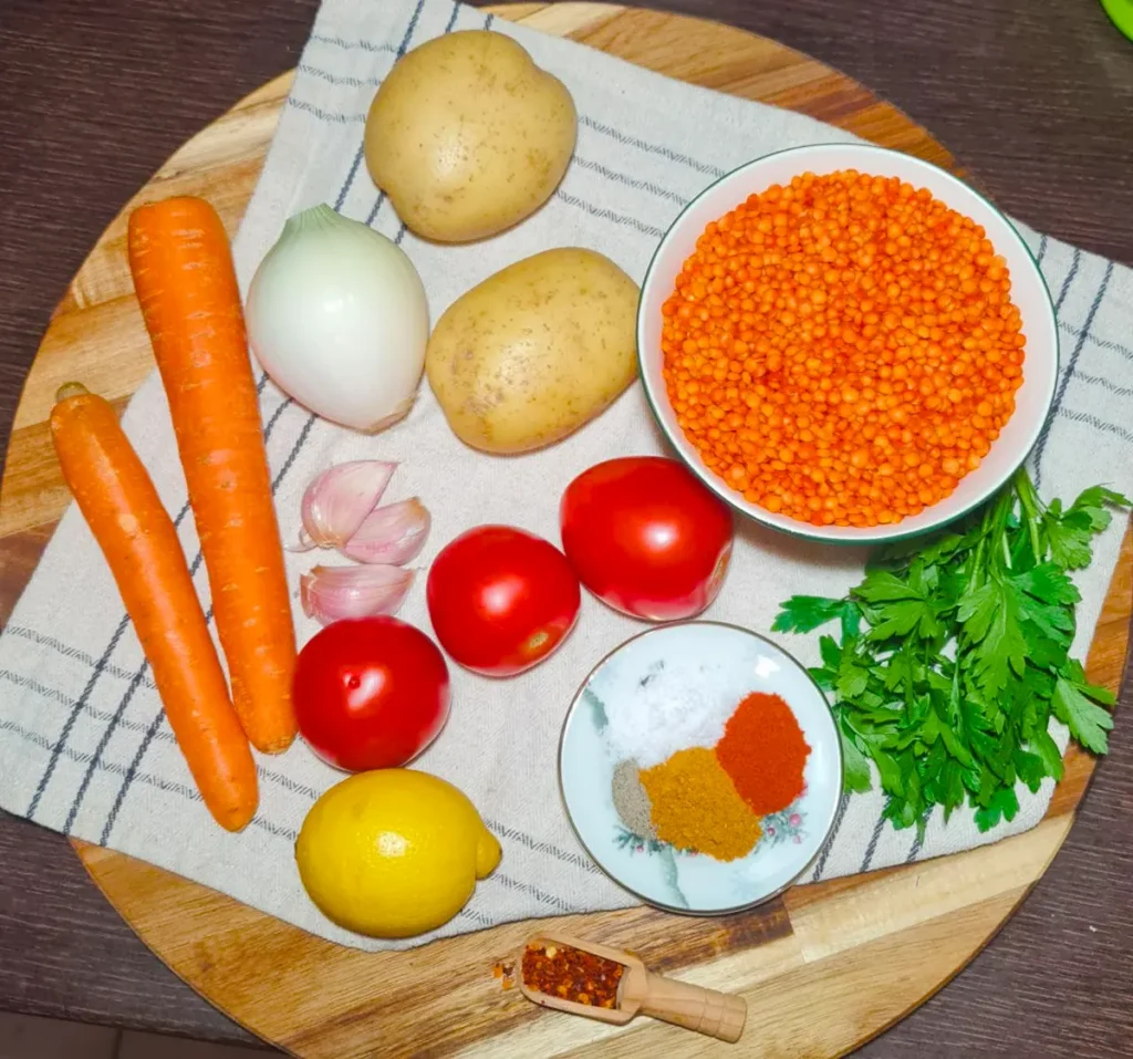Flat lay of raw ingredients for Turkish lentil soup on a wooden board, including a bowl of red lentils, carrots, potatoes, tomatoes, onion, garlic, lemon, parsley, and spices.