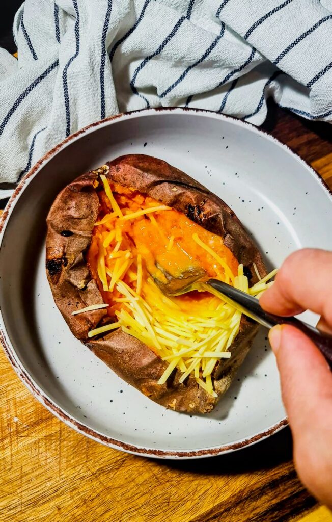 A hand using a fork to mash the inside of a baked sweet potato with vegan cheese to make vegan stuffed sweet potatoes.