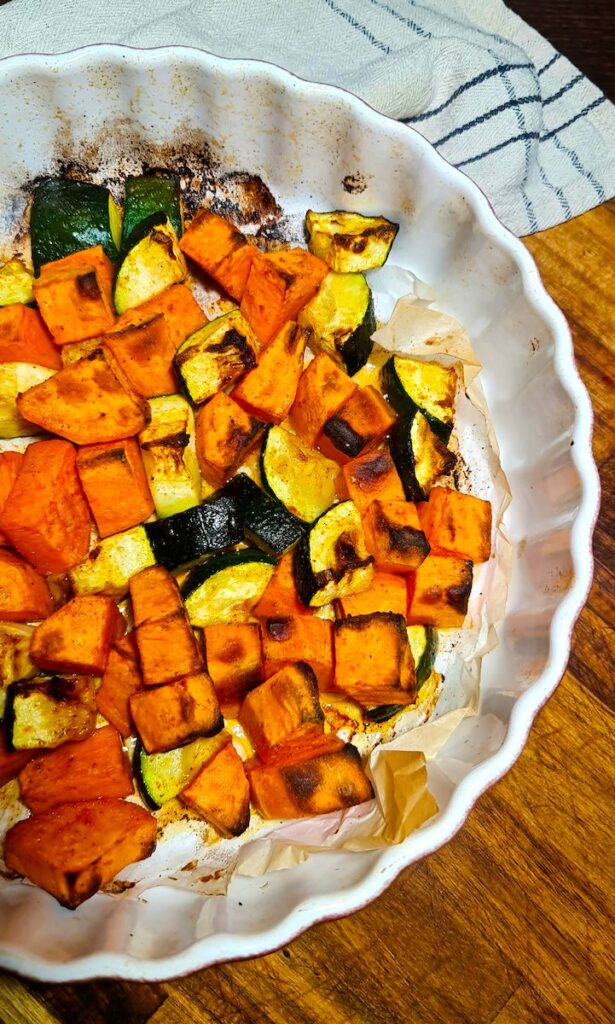 Golden, slightly charred roasted sweet potatoes and zucchini chunks in a white dish for a roasted vegetable quinoa bowl.