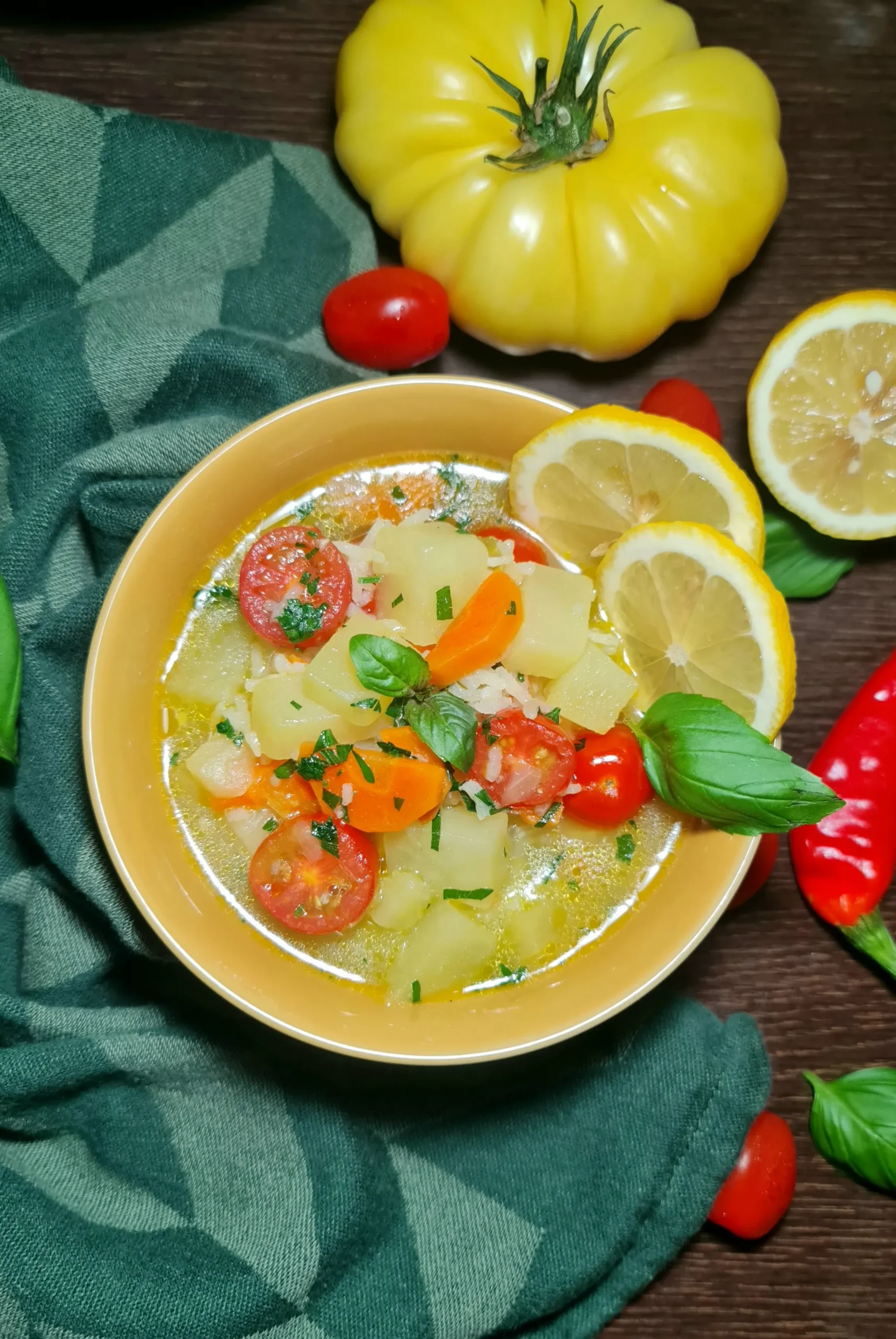 A rustic table setting featuring a yellow bowl of potato rice soup surrounded by fresh ingredients like lemon, basil, and a yellow tomato.