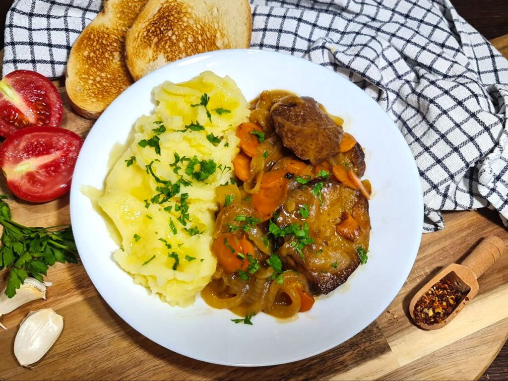 A rustic tabletop presentation of savory seitan with wine sauce, paired with creamy mashed potatoes, fresh tomatoes, and crisp toast on a wooden board.
