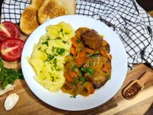 A rustic tabletop presentation of savory seitan with wine sauce, paired with creamy mashed potatoes, fresh tomatoes, and crisp toast on a wooden board.