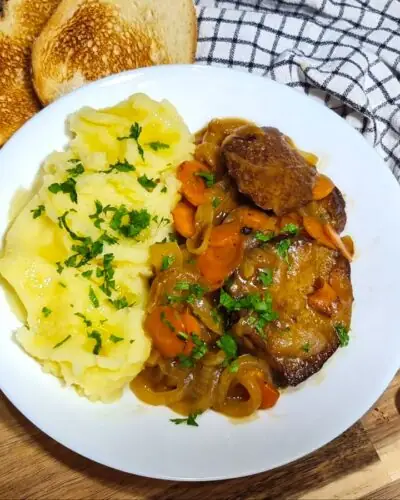 A rustic tabletop presentation of savory seitan with wine sauce, paired with creamy mashed potatoes, fresh tomatoes, and crisp toast on a wooden board.