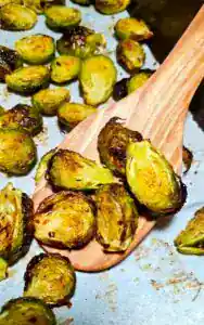A wooden spatula lifting crispy caramelized roasted brussels sprouts off a parchment-lined baking pan.