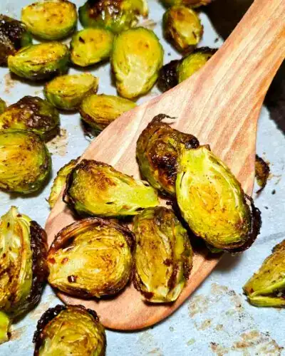 A wooden spatula lifting crispy caramelized roasted brussels sprouts off a parchment-lined baking pan.