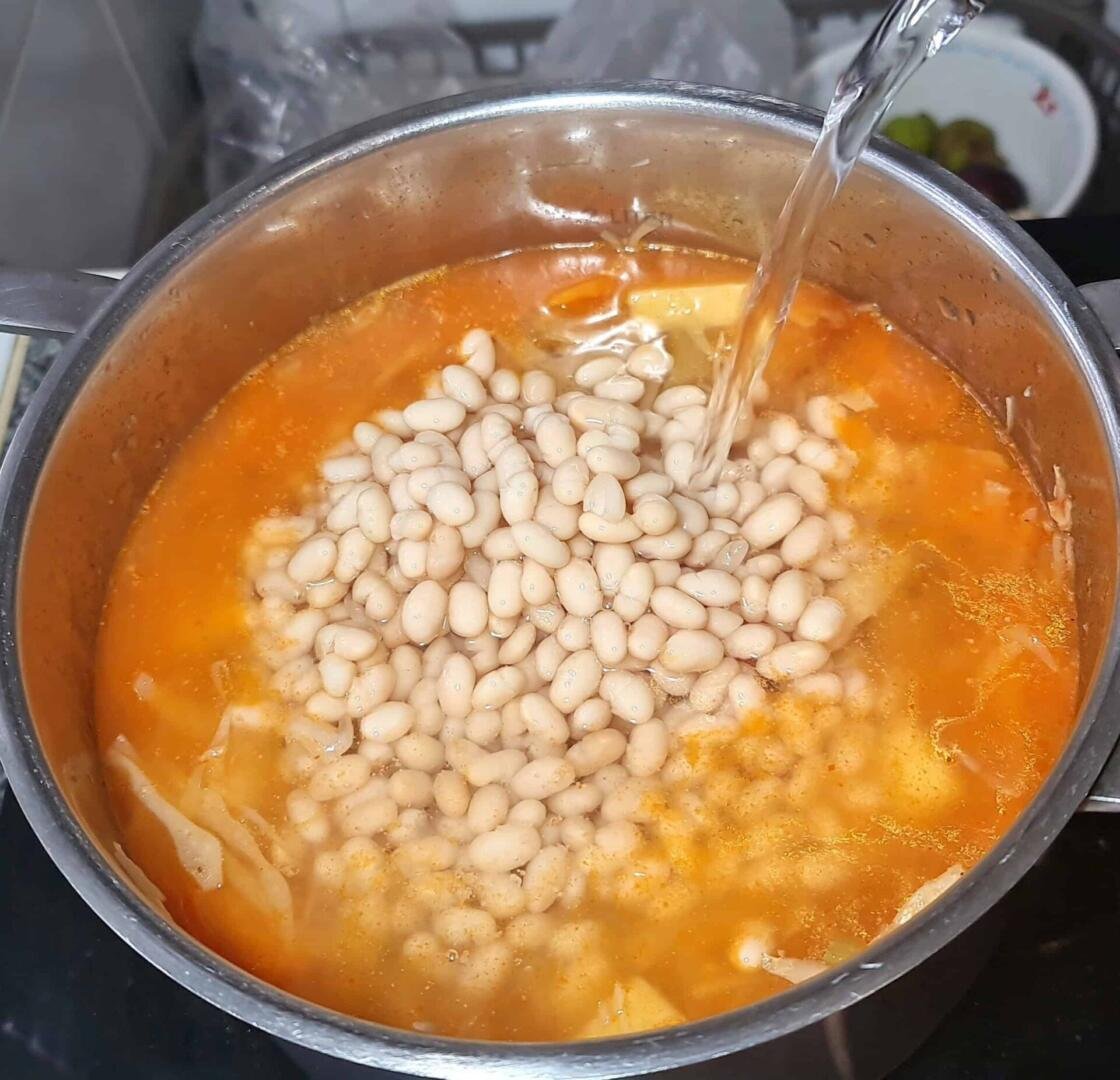 A pot of cabbage soup with white beans being added, followed by a stream of clear water to create the broth.
