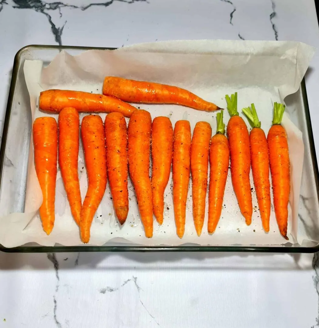 Whole fresh carrots seasoned with salt and pepper, lined up on parchment paper in a glass baking dish and ready to be roasted.