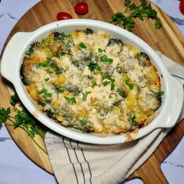 A freshly baked vegan broccoli potato casserole in a white dish, with a golden-brown, crispy topping and a spoon taking a scoop out to show the creamy interior.