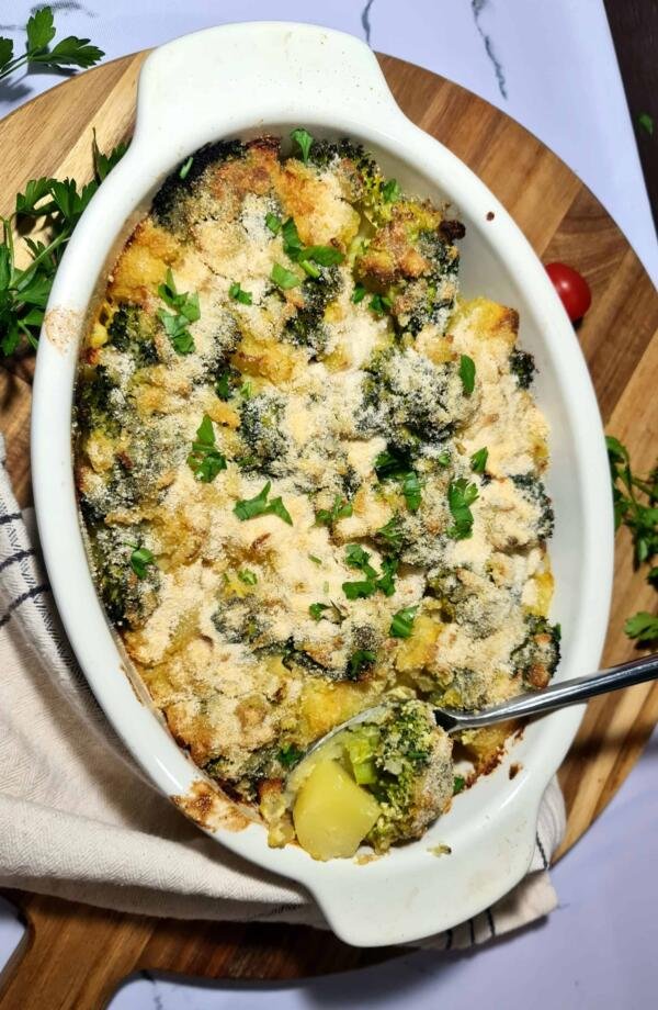 A freshly baked vegan broccoli potato casserole in a white dish, with a golden-brown, crispy topping and a spoon taking a scoop out to show the creamy interior.