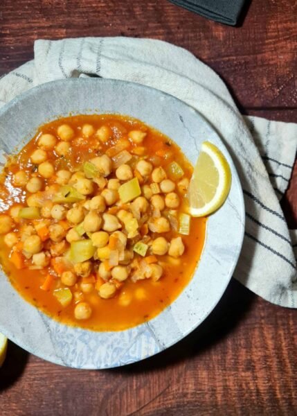A rustic blue and white bowl filled with hearty chickpea and zucchini soup in a tomato broth, garnished with a lemon wedge and set on a dark wood table.