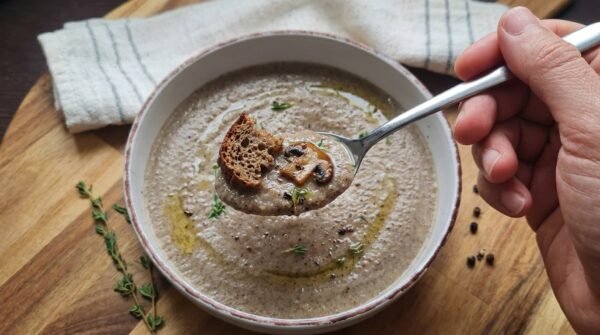A close-up of a spoon held over a bowl of soup, containing creamy mushroom soup topped with a small piece of toasted bread and a sautéed mushroom slice.