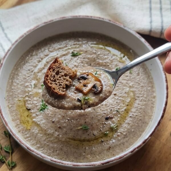 A close-up of a spoon held over a bowl of soup, containing creamy mushroom soup topped with a small piece of toasted bread and a sautéed mushroom slice.