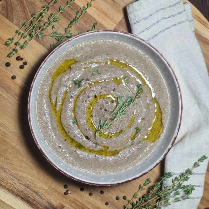 A bowl of creamy mushroom soup beautifully garnished with an olive oil swirl and a fresh sprig of thyme on a wooden cutting board.
