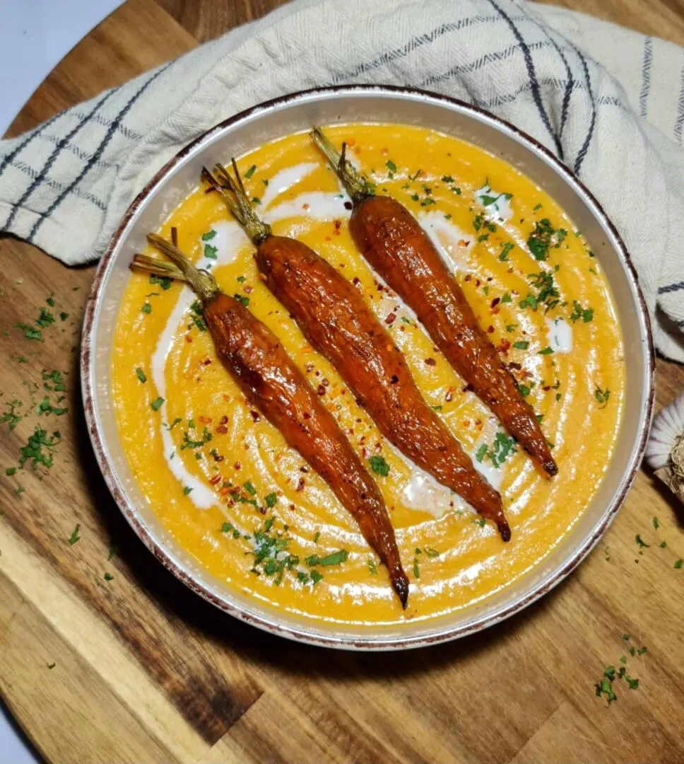 A rustic bowl of creamy orange roasted carrot potato soup, elegantly garnished with three whole roasted carrots, a swirl of coconut cream, and fresh parsley, set on a wooden board.