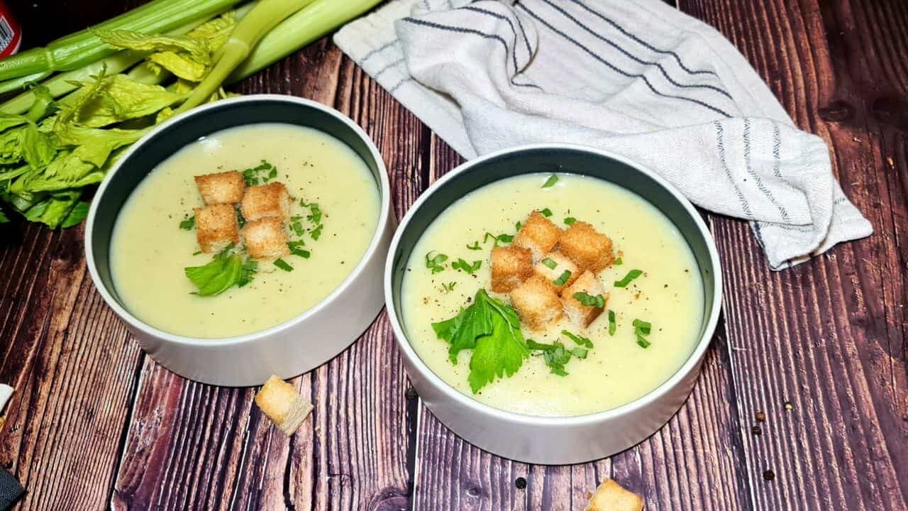 A wide shot of two bowls of creamy potato celery soup on a dark wood table, garnished with croutons and fresh parsley. A fresh stalk of celery and a kitchen towel are in the background.