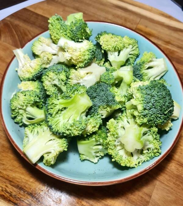 A light blue bowl filled with fresh, bright green broccoli florets, cut into bite-sized pieces and sitting on a wooden cutting board.