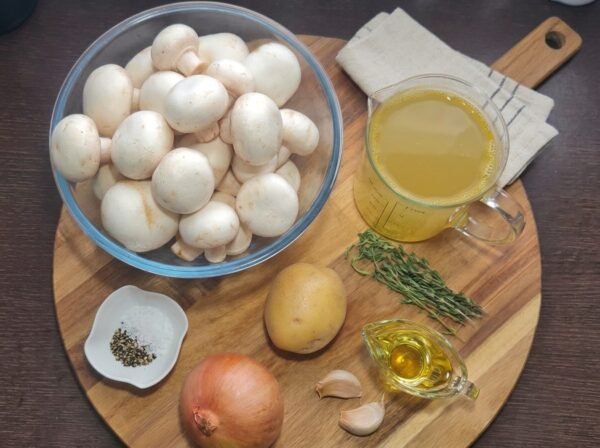 Flat lay of creamy mushroom soup ingredients including fresh white mushrooms, a potato, an onion, garlic, broth, olive oil, and herbs on a wooden board.