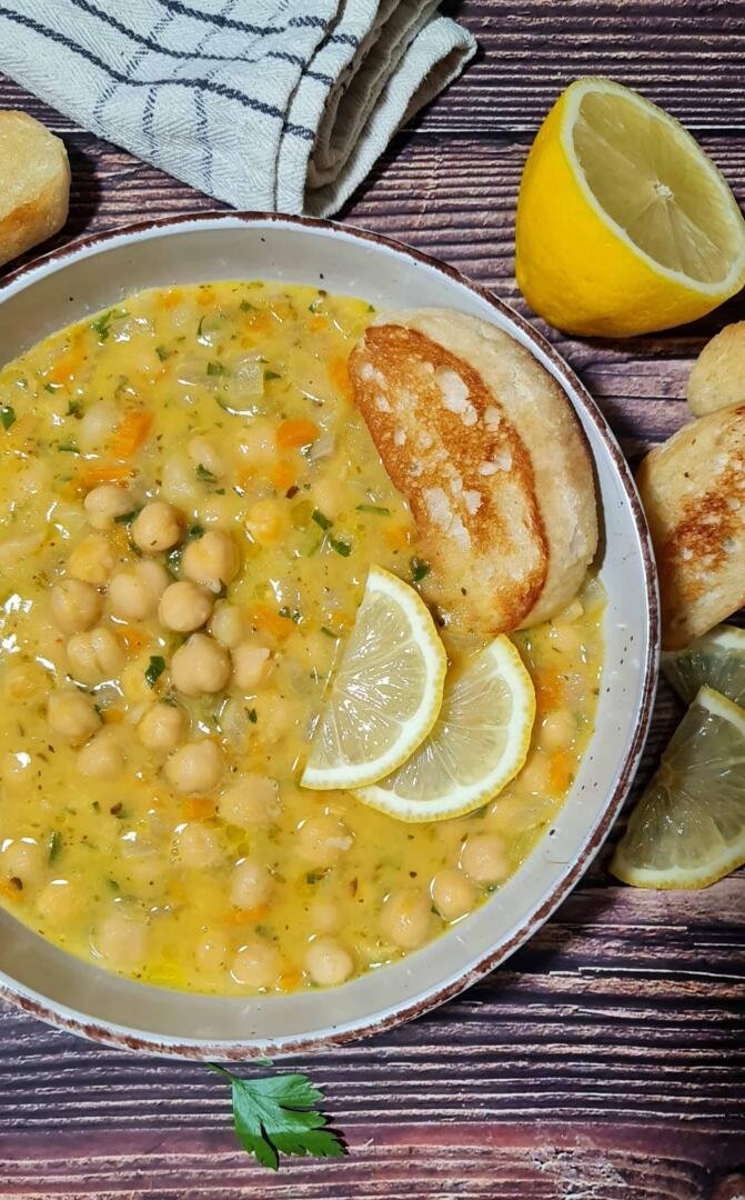 A rustic bowl of creamy Greek chickpea soup, garnished with fresh lemon slices and a piece of toasted bread, set on a dark wooden background.