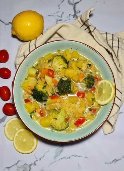 A light blue bowl filled with hearty autumn vegetable soup, showing chunks of potato, broccoli, rice, and carrots, garnished with a fresh lemon wedge on a marble surface.
