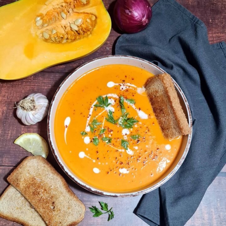 A rustic bowl of creamy roasted winter vegetable soup, garnished with fresh parsley, coconut cream, and chili flakes, served with toasted bread on a dark wood table.