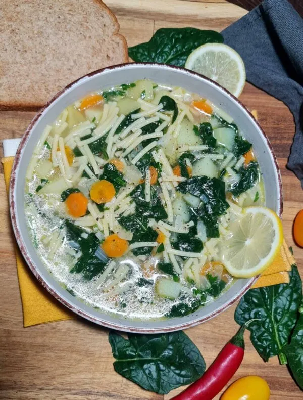 A rustic bowl of clear vegan spinach noodle soup with small noodles, potatoes, and spinach, garnished with lemon and served with a slice of bread on a wooden surface.