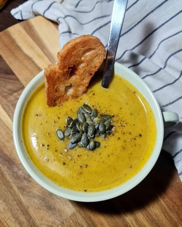 Creamy pumpkin zucchini soup topped with pumpkin seeds, served with toasted bread on a wooden board.