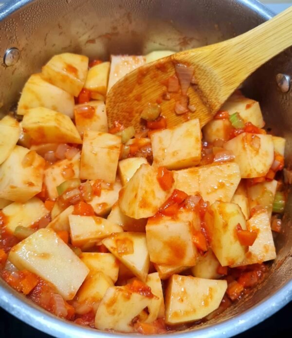 Large cubes of potato being stirred into the sautéed vegetable and tomato base in the pot, an essential step in making the thick potato and peas soup.