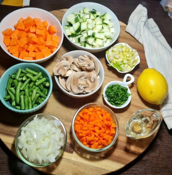 Chopped ingredients for easy vegetable soup: sweet potato, zucchini, green beans, mushrooms, onion, carrot, leek, parsley, lemon, and oil on a wooden board.