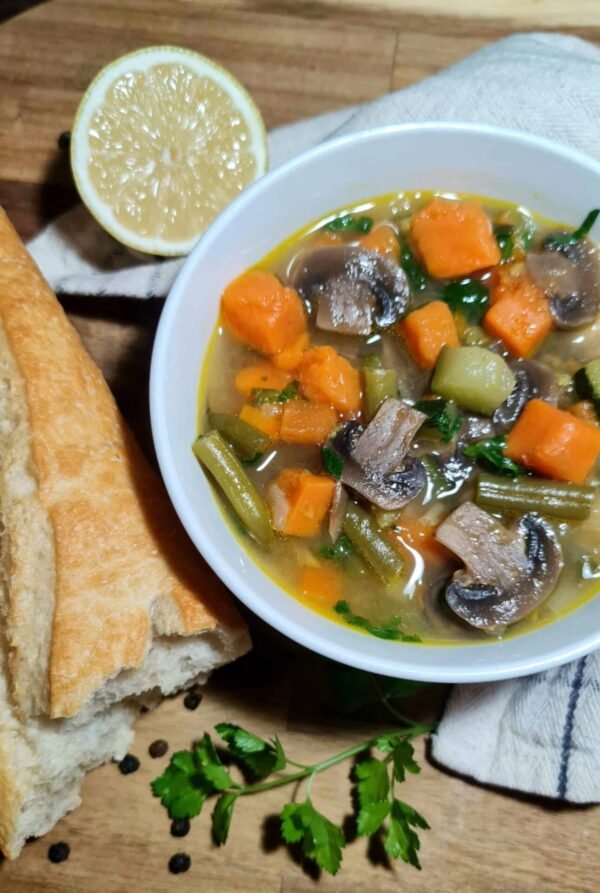 Served bowl of easy vegetable soup with tender mushrooms, sweet potato, zucchini, and green beans, lemon wedge and bread nearby.