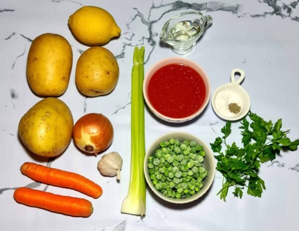 All ingredients for the vegan potato and peas soup laid out: potatoes, carrots, onion, celery, frozen peas, tomato sauce, lemon, garlic, and seasoning on a marble surface.