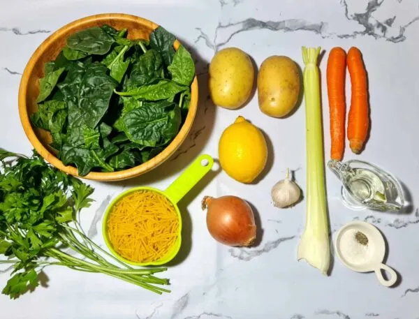 Whole ingredients, including spinach, potatoes, noodles, carrots, celery, onion, lemon, and garlic, laid out on a white marble background for spinach noodle soup.