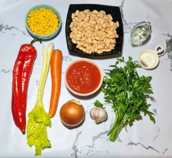 All ingredients needed for the vegan bean and corn soup, including white beans, corn, red pepper, onion, celery, garlic, parsley, tomato sauce, and oil, laid out on a marble surface.