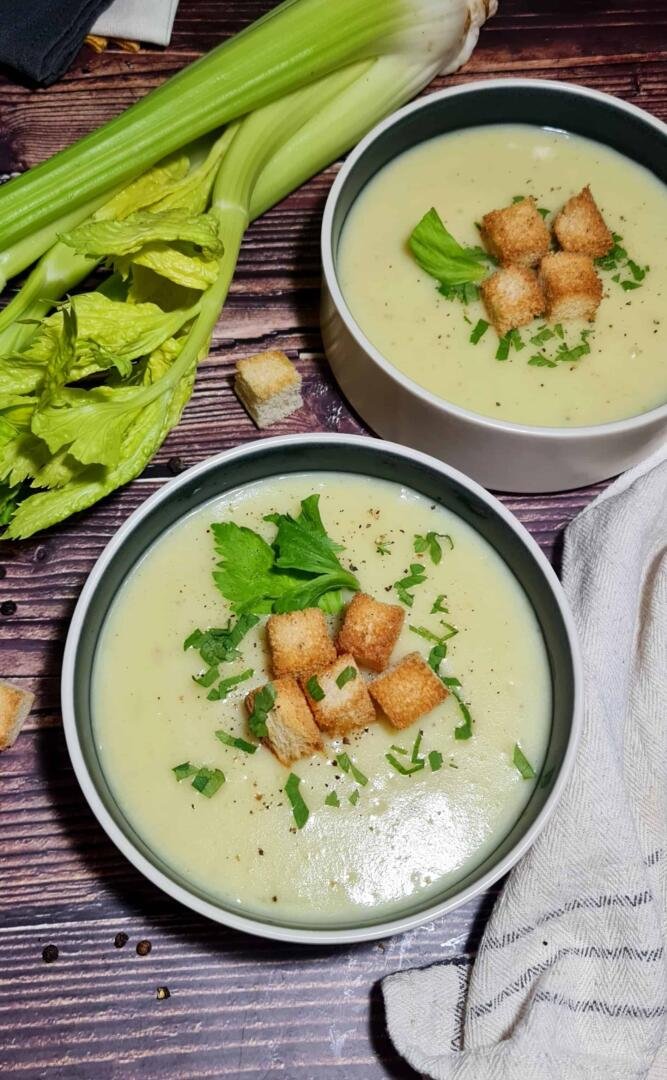 Two rustic bowls of creamy, light green potato celery soup, garnished with golden-brown croutons and fresh parsley, set on a dark wood table.